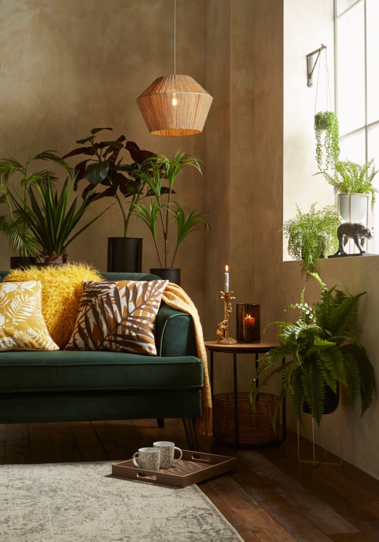 Deep green sofa surrounded by fresh plants, soft palm print cushions and a neutral blanket. All from Matalan.