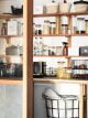 White walled pantry with wooden shelves, filled with glass jars full of food. Dotted around are black wire baskets and cleaning products.