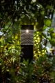 Black metal solar bollard light with cut-out holes shining a pattern onto nearby plants.