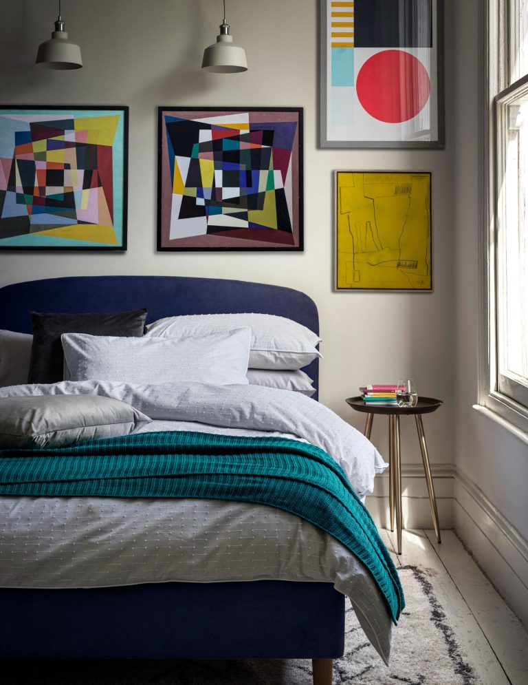 Navy fabric Mila bed with bright coloured artwork, light blue repeat texture stripe bedset, pure cotton knitted throw and mango and brass side table by Marks and Spencer.
