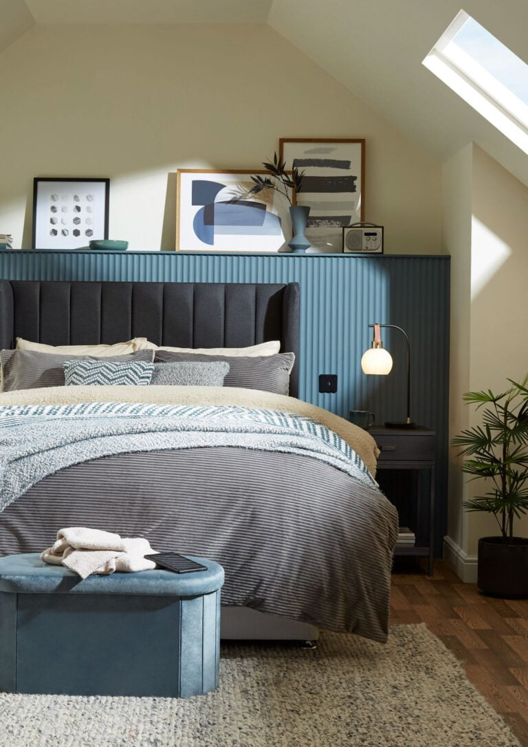 A bedroom with a bed, nightstands, and a plant. The bed is made up with a gray comforter and a blue blanket. The nightstands are next to the bed and have a lamp on them. There is a plant in the corner of the room. The wall behind the bed has a contemporary modern urban designer bedroom size headboard.