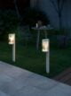 The image shows two Nordlux Coupar garden lamps in the grass at night. The lamps are made of white metal and have a cylindrical shape. They are illuminated and are casting a soft light on the grass. The image is set at night and the sky is dark. The background is a garden with trees and flowers.