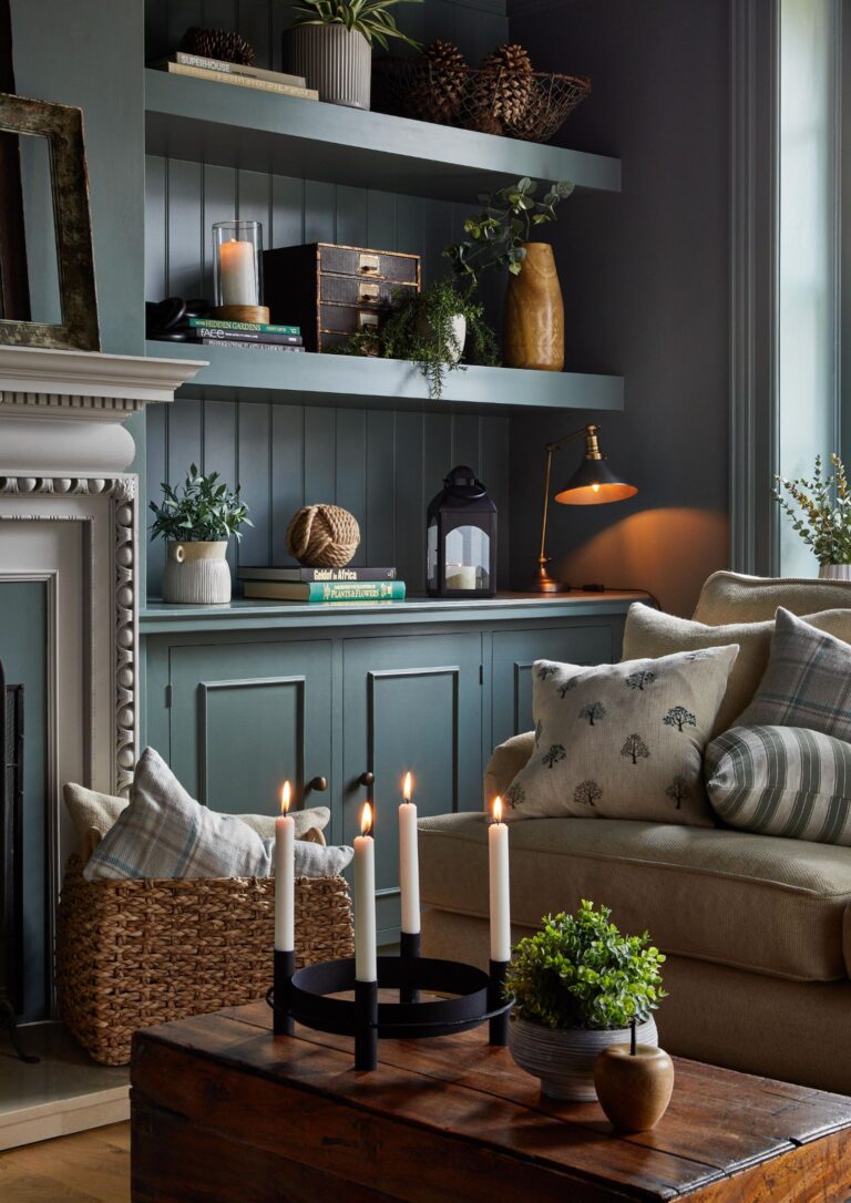 The image shows a cozy living room with a brown couch, a coffee table with candles on it, and shelves on the walls. There is a fireplace in the background. The shelving either side of the fireplace is a nice greeny-blue colour with natural light shining in.