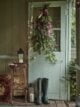 Christmas door wreath made with a bunch of greenery, wrapped in a ribbon, lights and lanterns beside the door.