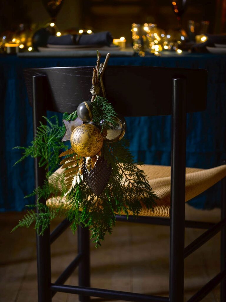 Christmas greenery with baubles hanging off the back of dining chair at a Christmas dinner table.