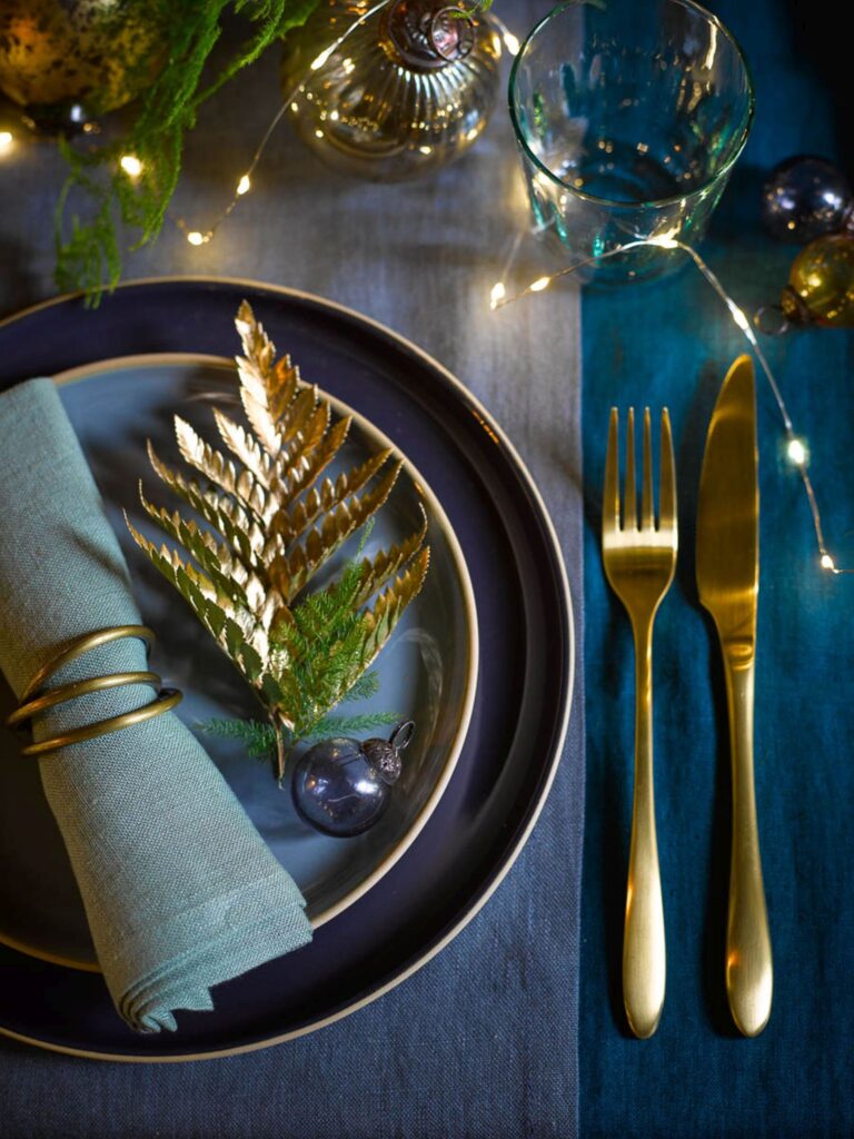 Brass cutlery, with gold Christmas baubles, warm white led lights and a dark blue table cloth by Garden Trading.