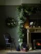 Green living room with a large Christmas garland hanging down the wall, beside a fireplace covered in candles, baubles and wire lights. Beside the fireplace is a spindle chair, paper Christmas trees and lanterns.