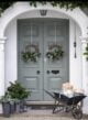 Light green front door with two wreaths, potted Christmas trees and a wheelbarrow full of presents.