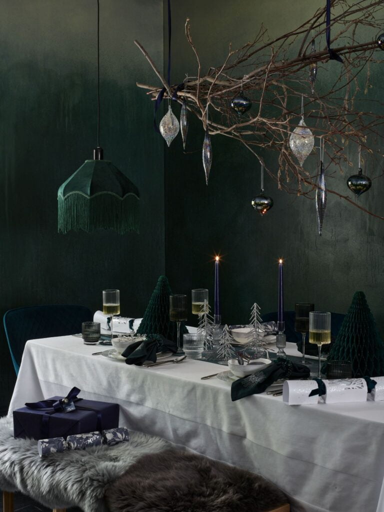 Dark and moody green Christmas dinner table backdrop, with hanging wooden branch covered in Christmas ornaments, white table cloth and green paper Christmas trees by Marks and Spencer.