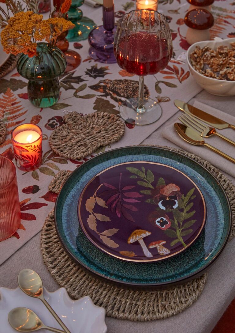 A close-up of a beautifully set table for a fall dinner party. A dark purple plate with a floral and mushroom design sits on a woven placemat. A glass of red wine, a candle, and a bowl of walnuts are also visible on the table.