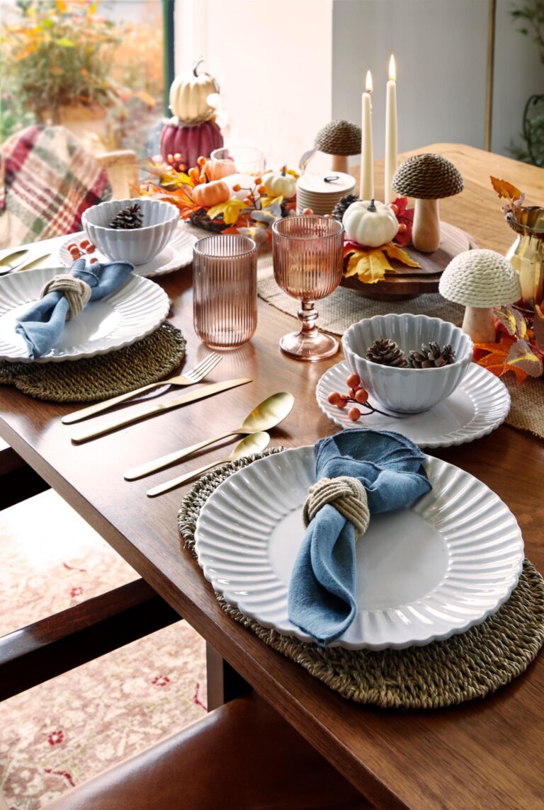 A table set for a fall dinner party. A wooden table is adorned with a white tablecloth and a centrepiece featuring pumpkins, mushrooms, and autumn leaves. Plates, bowls, and glasses are arranged around the table, and napkins are tied with raffia. Candles and decorative mushrooms add to the fall ambiance.