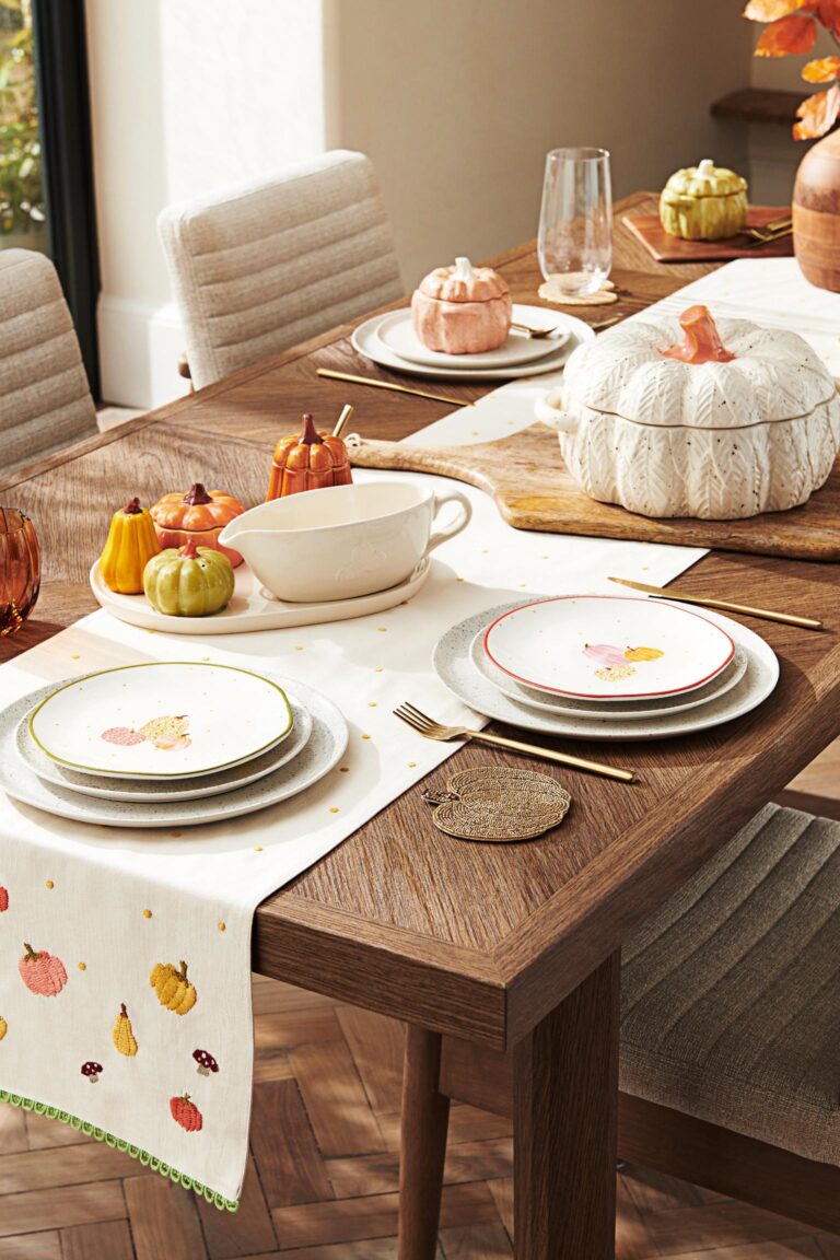 A wooden dining table set for a fall feast. The table is adorned with a white tablecloth featuring a pumpkin and mushroom pattern. A pumpkin-shaped casserole dish sits at the centre, surrounded by smaller pumpkin-shaped bowls and pitchers. Plates, napkins, and cutlery are arranged around the table, and a vase of fall flowers adds a touch of autumnal colour.