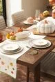 A wooden dining table set for a fall feast. The table is adorned with a white tablecloth featuring a pumpkin and mushroom pattern. A pumpkin-shaped casserole dish sits at the centre, surrounded by smaller pumpkin-shaped bowls and pitchers. Plates, napkins, and cutlery are arranged around the table, and a vase of fall flowers adds a touch of autumnal colour.