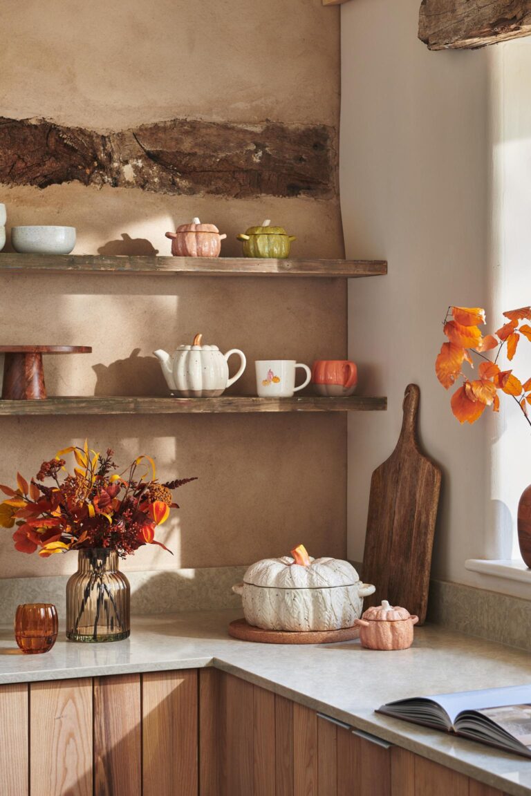 Kitchen with autumnal vibes. There are shelves filled with pumpkin-shaped dishes, a vase of fall flowers, and a wooden cutting board.