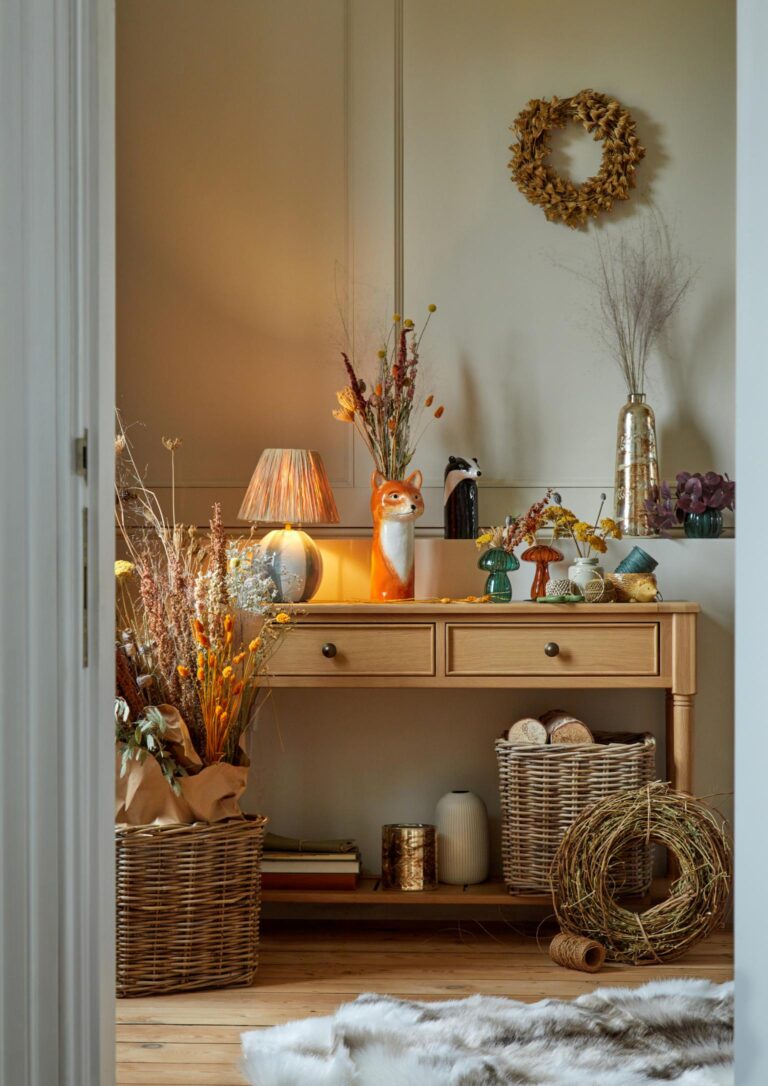 Hallway decorated for autumn. A wooden console table holds a variety of autumnal decor, including a vase of dried flowers, a ceramic fox figurine from Dunelm. Wicker baskets and a woven wreath complete the autumnal ambiance.