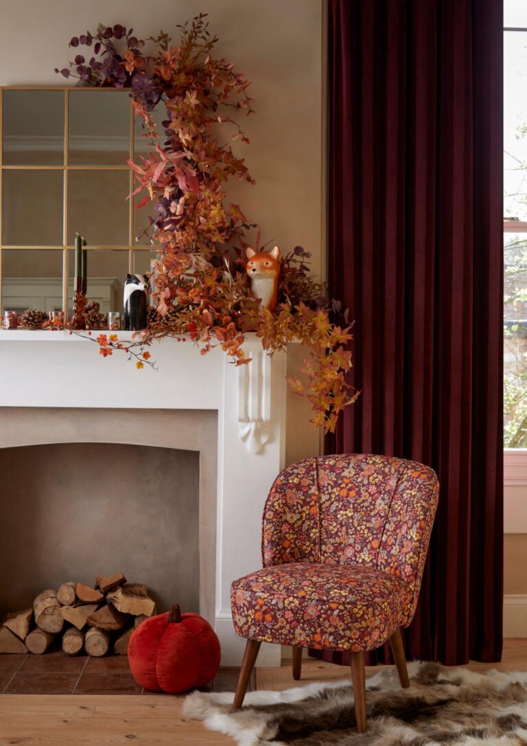 Cosy living room with a fireplace decorated with autumn leaves and a cute fox figurine. A comfy armchair with a floral pattern sits nearby, and a stack of firewood is piled beside it. A big red curtain hangs in the background, and a mirror reflects the autumnal vibe.