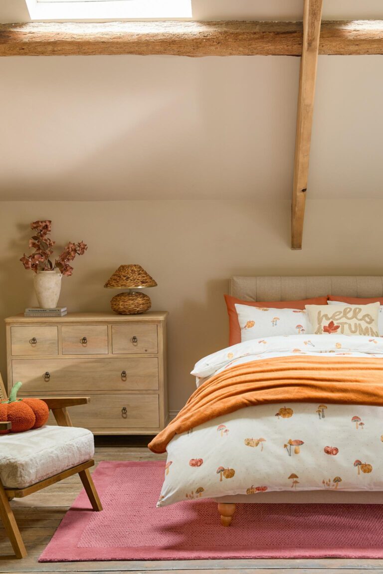 An autumn bedroom with a white duvet cover featuring a pattern of mushrooms and pumpkins in autumnal colours from Next. The bed is made up with an orange throw blanket and a pillow that says "Hello Autumn."