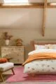An autumn bedroom with a white duvet cover featuring a pattern of mushrooms and pumpkins in autumnal colours from Next. The bed is made up with an orange throw blanket and a pillow that says "Hello Autumn."