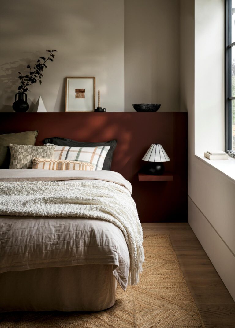 A cosy bedroom with a white duvet cover and a textured throw from Marks and Spencer. The bed is set against a wall with a deep red accent. A small bedside table with a lamp sits next to the bed.