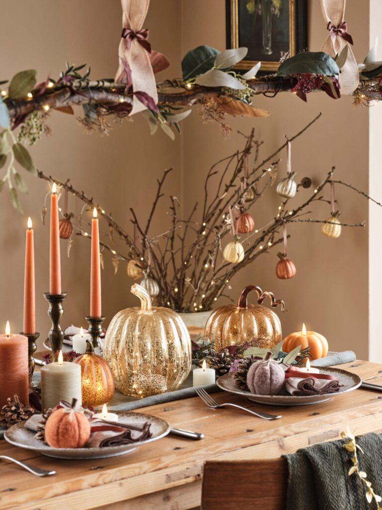Dining room table set for an autumn dinner party. The table is adorned with a runner and plates, and there are several pumpkins scattered throughout the space. Candlesticks hold tall, orange candles, creating a warm and inviting atmosphere. A branch with hanging decorations and fairy lights hangs above the table.
