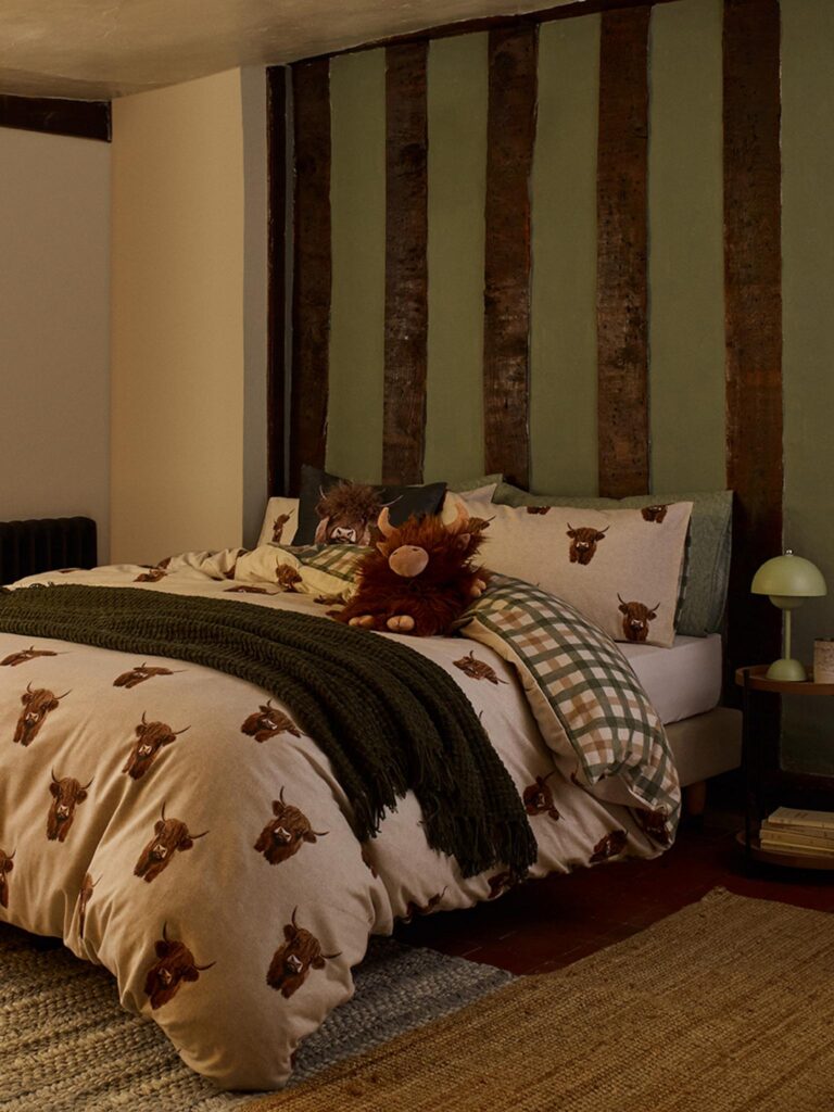 A cosy bedroom with a bed featuring a duvet cover and pillowcases with a pattern of Highland cows by George Home at Asda. A green throw blanket is draped over the bed, and a stuffed Highland cow toy sits on the pillow.