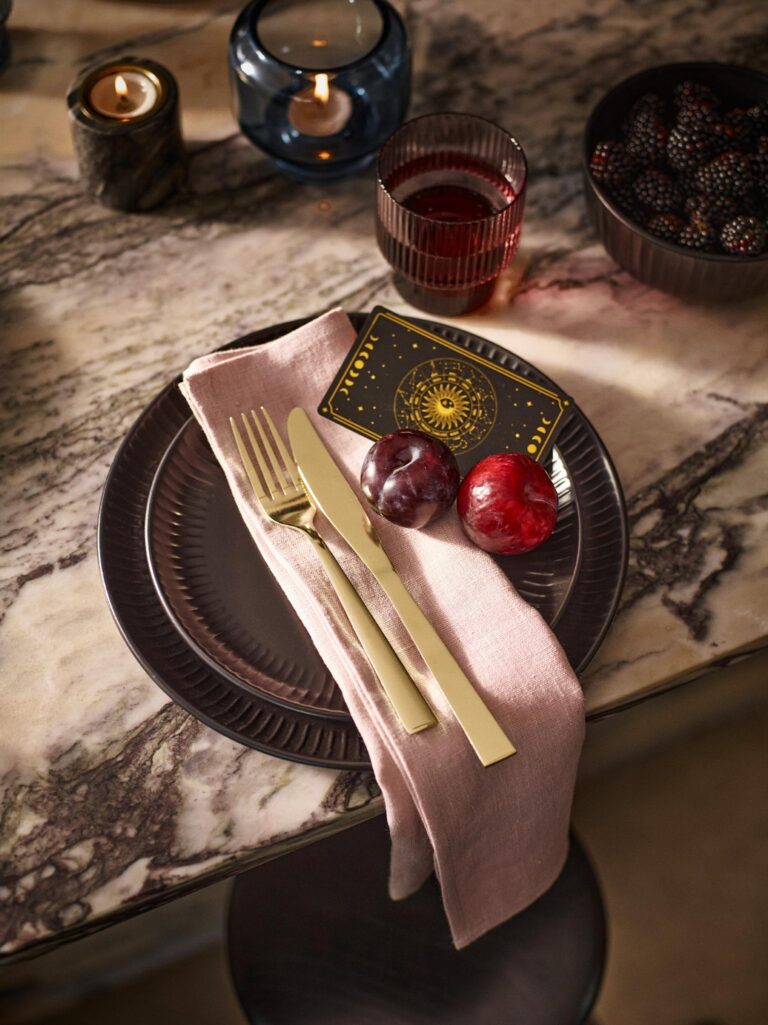 A dark plate set on a marble table. A pink napkin is folded next to a fork and knife, with two plums and a tarot card on top.