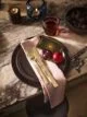 A dark plate set on a marble table. A pink napkin is folded next to a fork and knife, with two plums and a tarot card on top.