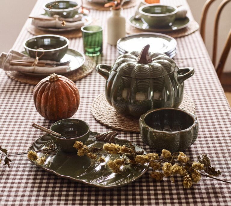 A table set for a fall dinner party. A large, green pumpkin-shaped casserole dish sits at the centre, surrounded by smaller pumpkin-shaped bowls and plates.