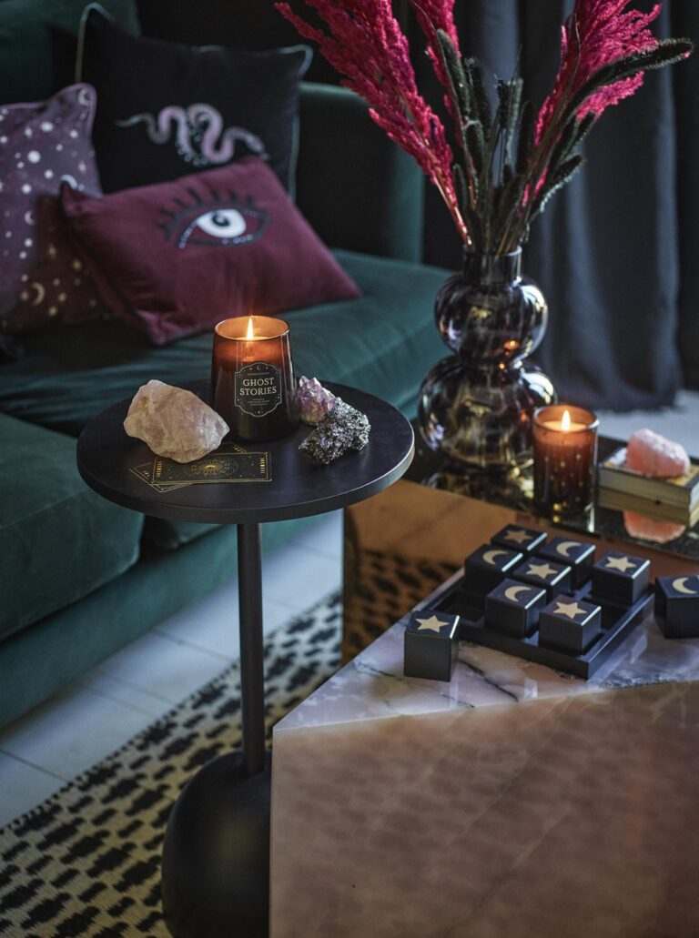 Autumn living room with a dark green sofa adorned with Halloween-themed pillows, including ones with ghosts and eyes from George Home. A black side table holds a lit candle labeled "Ghost Stories," surrounded by crystals and tarot cards. A larger coffee table features a black tray with small boxes decorated with stars and moons. A vase of dried flowers and another lit candle sit on the coffee table.