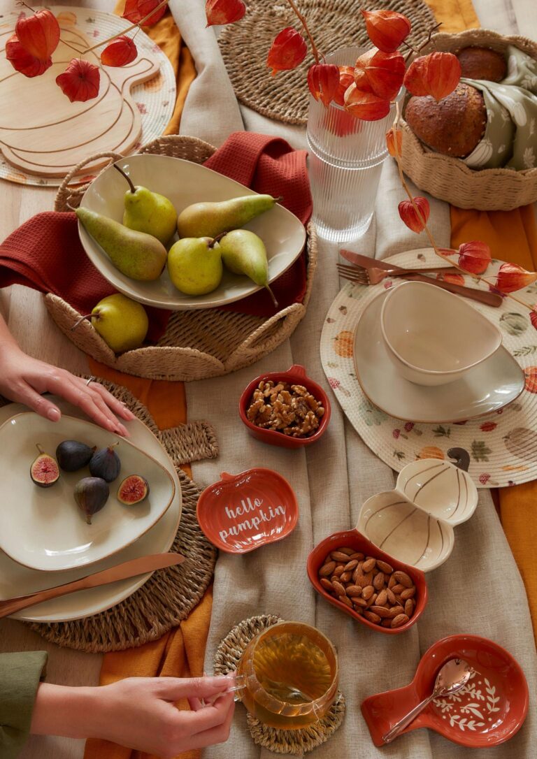 A cosy autumnal table setting with pumpkins, pears, and fall-themed dishes. The table is covered in a warm orange cloth with woven placemats.