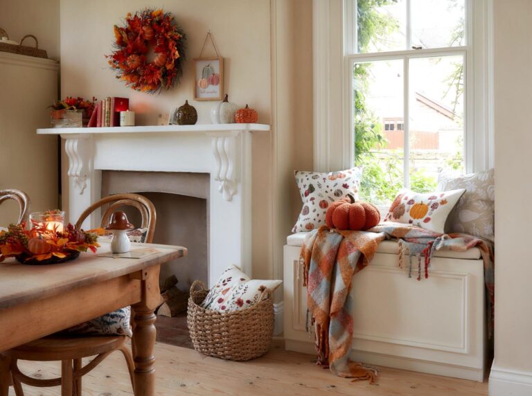 Autumn living room with a white fireplace decorated with autumnal decor, including a wreath, pumpkins, and candles from Matalan. A window seat is adorned with fall-themed pillows and a plaid throw blanket. A wooden dining table is set with autumn-inspired decorations, including a centerpiece with pumpkins and leaves.