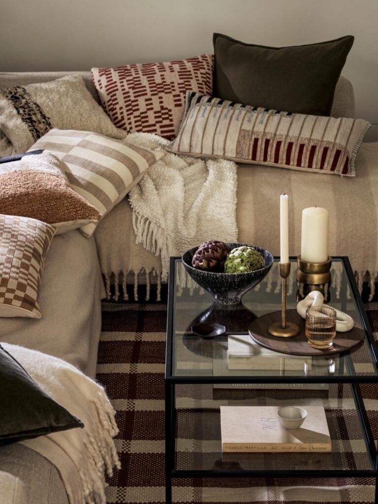 Living room with a sofa covered in a neutral throw blanket and cushions from Marks and Spencer. The sofa is adorned with a variety of colourful throw pillows, including patterned and textured options. A glass coffee table holds a bowl of decorative pinecones, a candle, and a book. A plaid rug covers the floor.