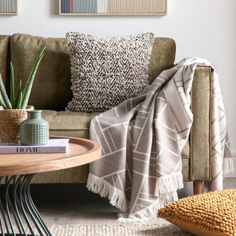 Cosy living room with a green sofa draped in a beige throw blanket with a geometric pattern from Very. A textured throw pillow sits on the sofa, and a coffee table holds a book and decorative vases.
