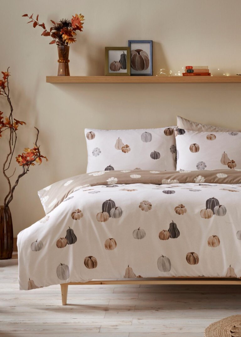 A bed with a white duvet cover featuring a pattern of pumpkins in various sizes and shades of orange, brown, and beige from Matalan. The bed is made up with beige pillowcases and is set against a neutral wall. A wooden shelf above the bed holds a vase of autumn leaves, framed pictures, and a string of fairy lights.
