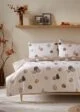 A bed with a white duvet cover featuring a pattern of pumpkins in various sizes and shades of orange, brown, and beige from Matalan. The bed is made up with beige pillowcases and is set against a neutral wall. A wooden shelf above the bed holds a vase of autumn leaves, framed pictures, and a string of fairy lights.