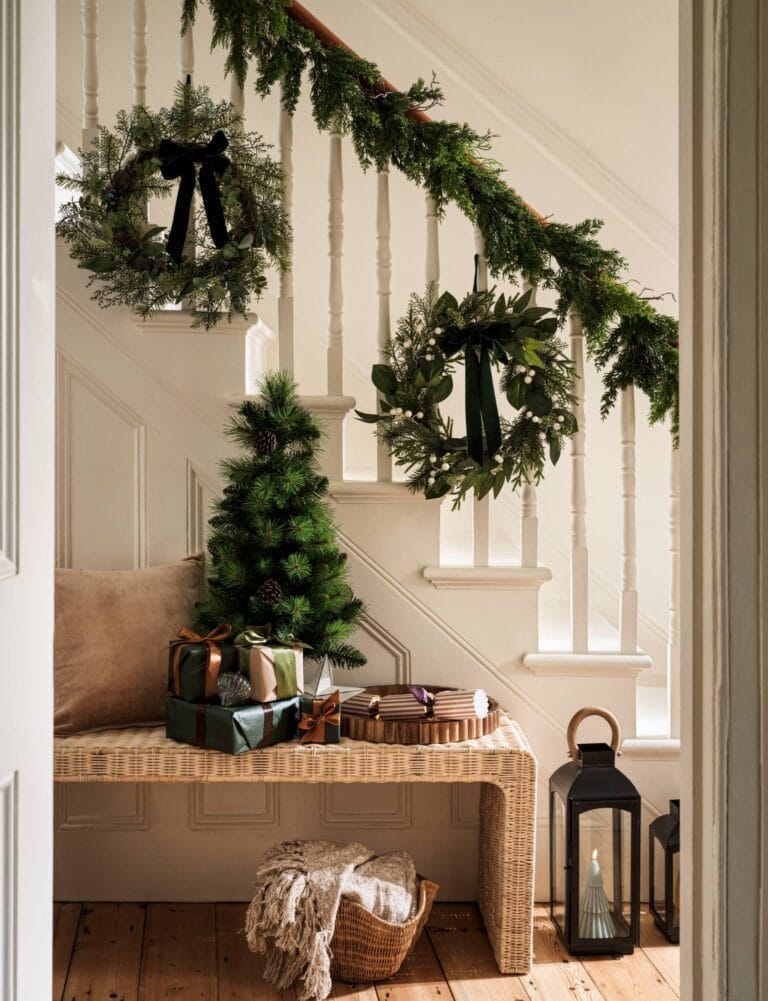 Staircase with a Habitat wreath trailing down the staircase and wreaths dotted along. A bench sits in the hallway with a Christmas tree, cracker and presents.