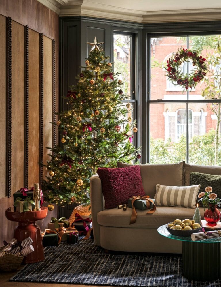Large bay window with a sofa sat inside, hanging Christmas wreath and a large Christmas tree. The Christmas style is very traditional with reds, golds and greens.