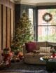 Large bay window with a sofa sat inside, hanging Christmas wreath and a large Christmas tree. The Christmas style is very traditional with reds, golds and greens.